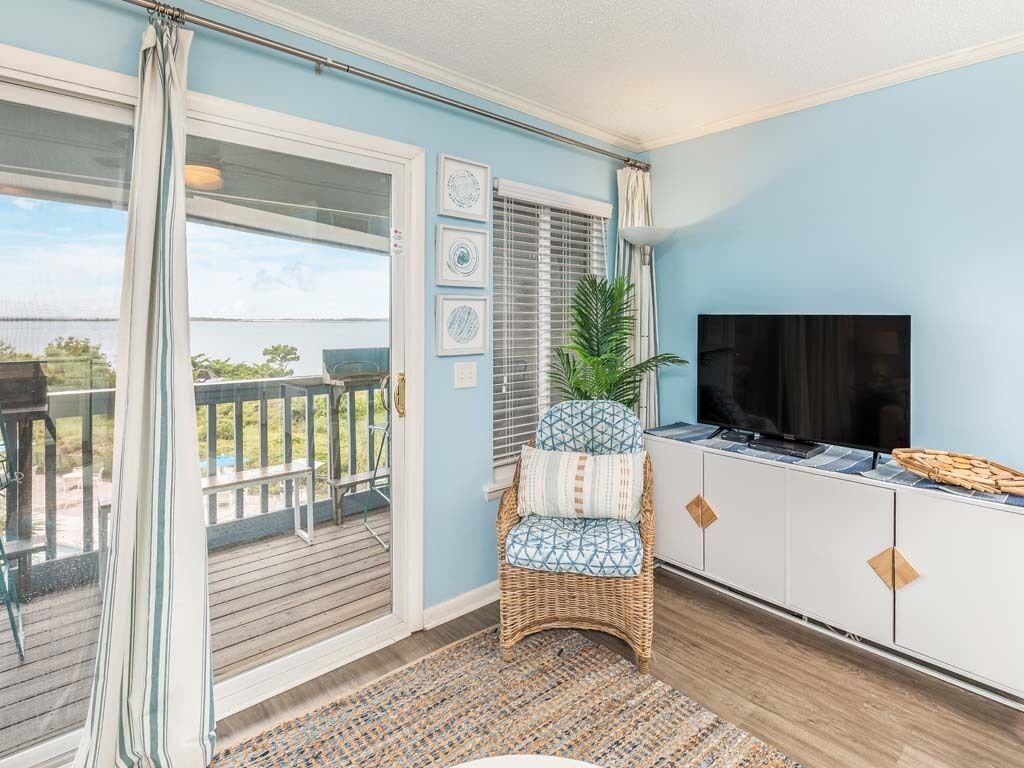 A room with a large sliding glass door leading to a balcony, a wicker chair with a cushion, a flat-screen TV on a white cabinet, and a patterned rug on the floor.