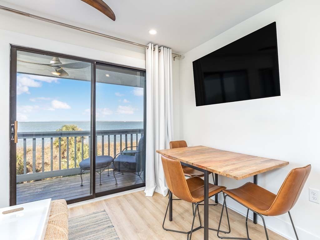 A room with a wooden dining table, two chairs, a TV mounted on the wall, and a large sliding glass door leading to a balcony with a view of the ocean.