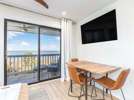 A room with a wooden dining table, two chairs, a TV mounted on the wall, and a large sliding glass door leading to a balcony with a view of the ocean.