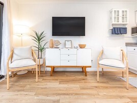 Una sala de estar con dos sillones blancos, un armario blanco, un televisor de pantalla plana y una planta en maceta.