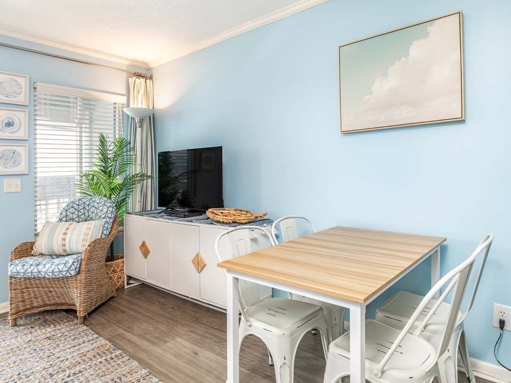 Une pièce avec un mur bleu clair, une table en bois avec des chaises blanches, un fauteuil en osier avec un coussin, une armoire blanche avec un téléviseur et un tableau encadré accroché au mur.