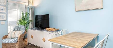 Een kamer met een lichtblauwe wand, een houten tafel met witte stoelen, een rieten stoel met kussen, een witte kast met een televisie en een ingelijste foto aan de muur.
