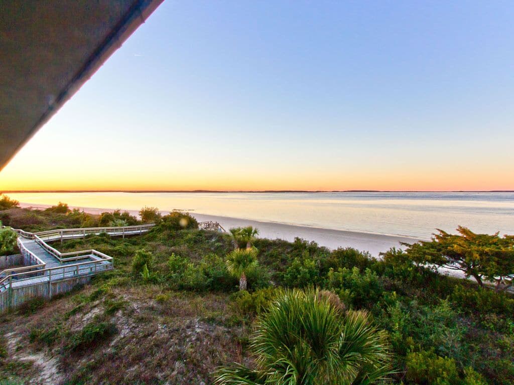 A coastal landscape with a wooden boardwalk leading to a beach, surrounded by greenery.