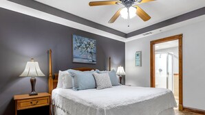 A bedroom with a large bed, two bedside tables with lamps, a ceiling fan, a painting on the wall, and a door leading to another room. - The Vue by Tybee Vacation Rentals (Tybee Island)
