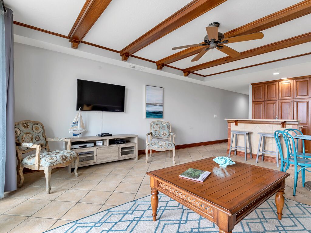 A spacious living room with a wooden ceiling, a flat-screen TV, a dining area with blue chairs, and a kitchen with wooden cabinets.