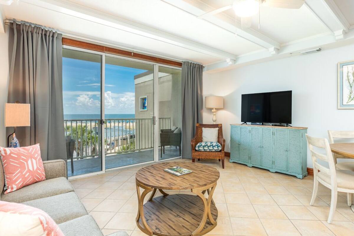 Sala de estar com sofá, mesa de madeira redonda, televisão e varanda com vista para a praia.