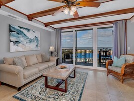 A spacious living room with a beige sofa, a coffee table, and a wicker chair. A large abstract painting adorns the wall, and a balcony with a view is visible through the glass doors.