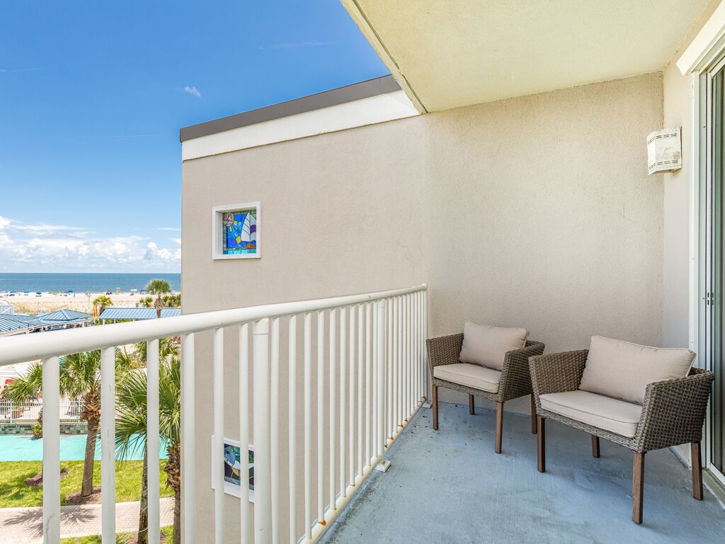 A balcony with two wicker chairs and a small table, overlooking a beach and pool area.