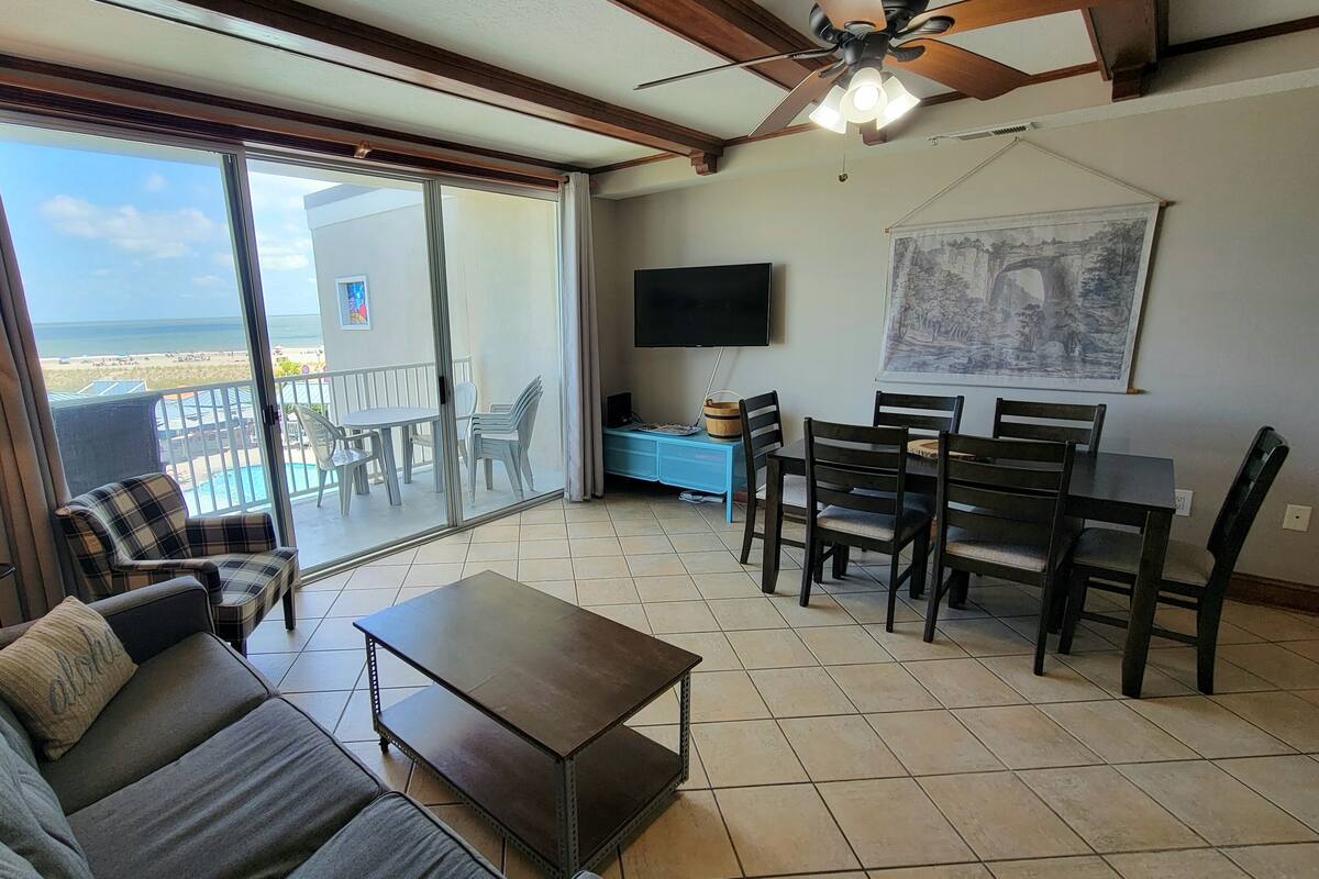 Sala de estar com sofá, mesa de centro, mesa de jantar e cadeiras. Há uma televisão na parede, um ventilador de teto e uma janela grande com vista para a praia.