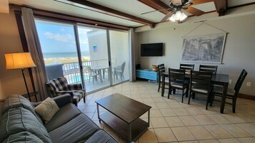 A living room with a sofa, coffee table, dining table, and chairs. There is a TV mounted on the wall, a ceiling fan, and a large window with a view of the beach.