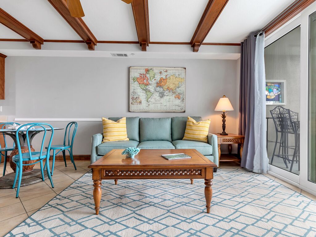 A living room with a wooden coffee table, a sofa with yellow and blue cushions, a dining table with blue chairs, and a map on the wall.