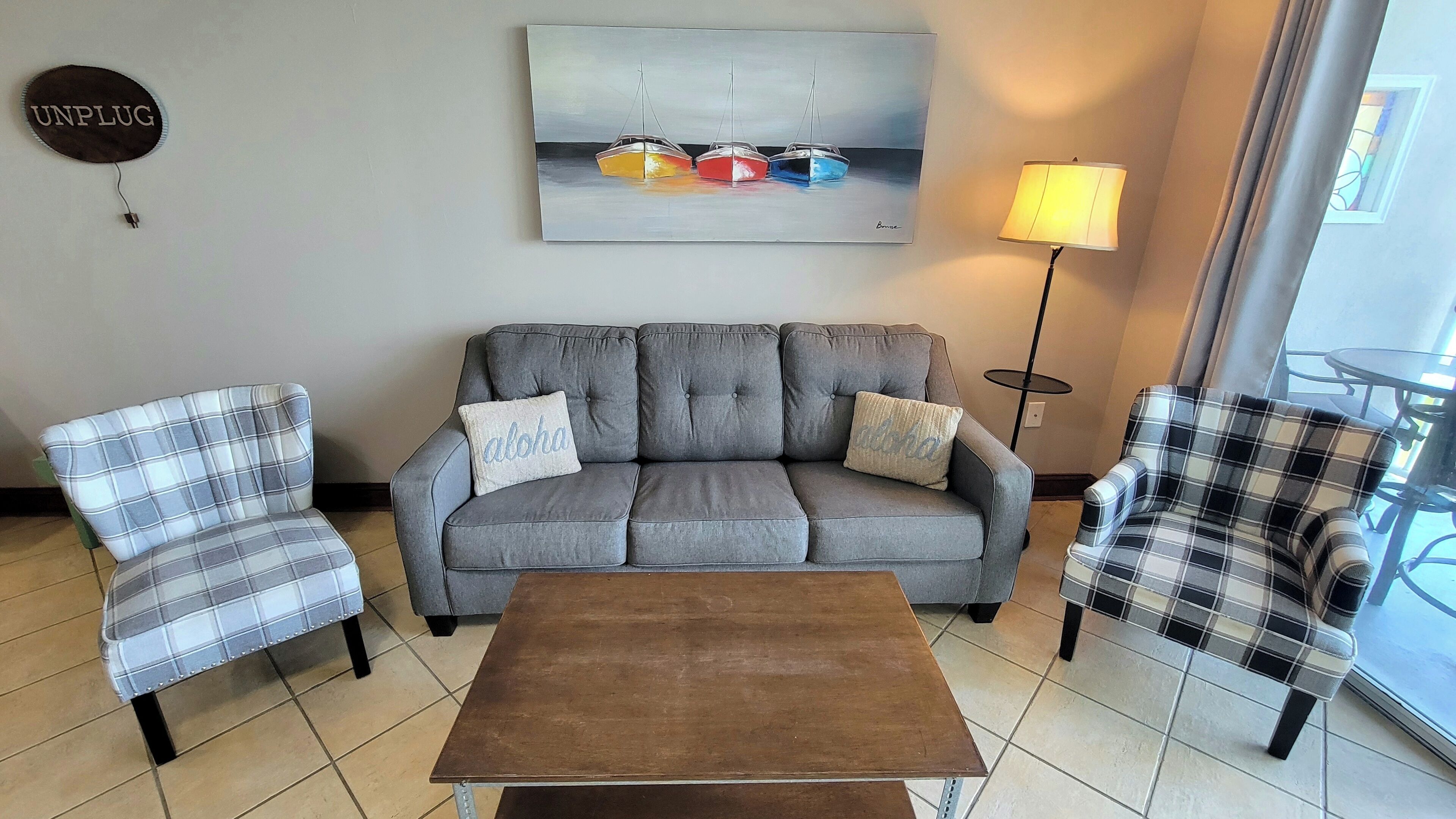 A living room with a grey sofa, two plaid armchairs, a wooden coffee table, a painting of boats, and a 'UNPLUG' sign.