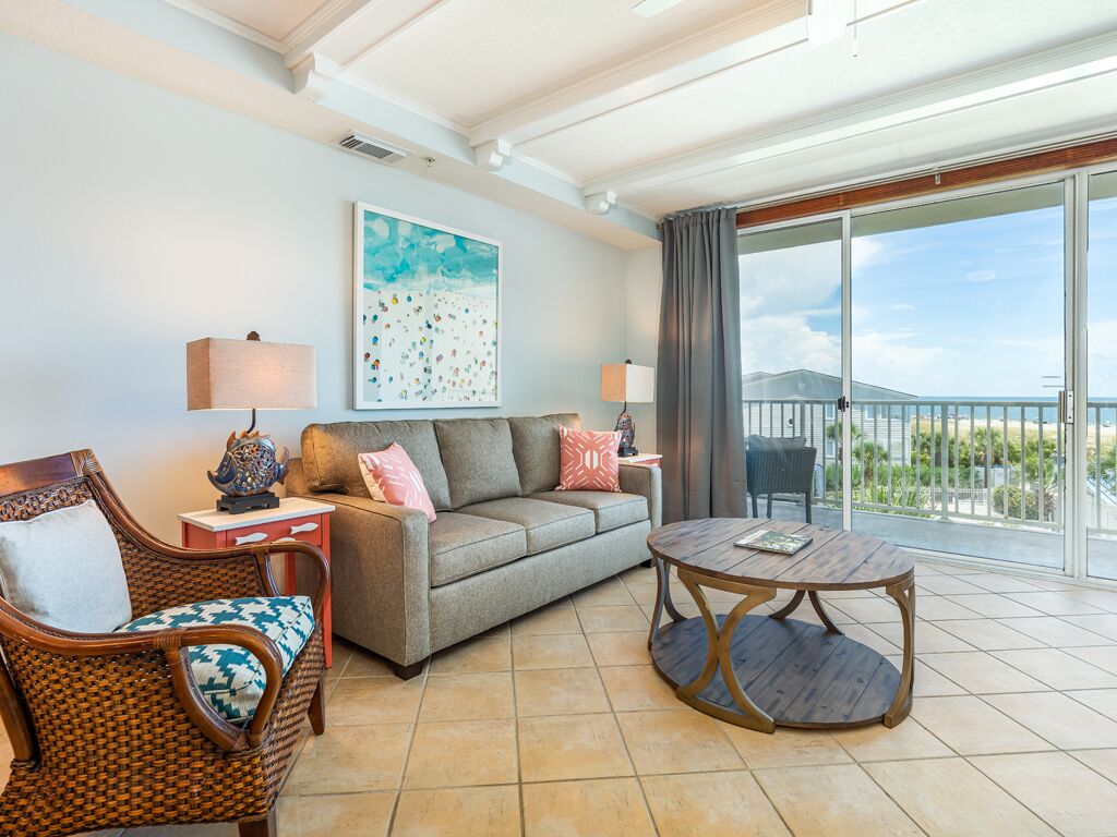 A living room with a sofa, a wicker chair, a round wooden table, and a balcony with a view.