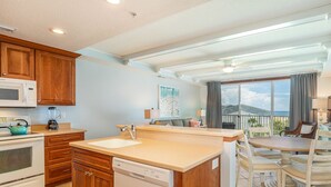 A modern kitchen with wooden cabinets, a dishwasher, and a large island. There is a dining area with a table and chairs. - The Vue by Tybee Vacation Rentals (Tybee Island)