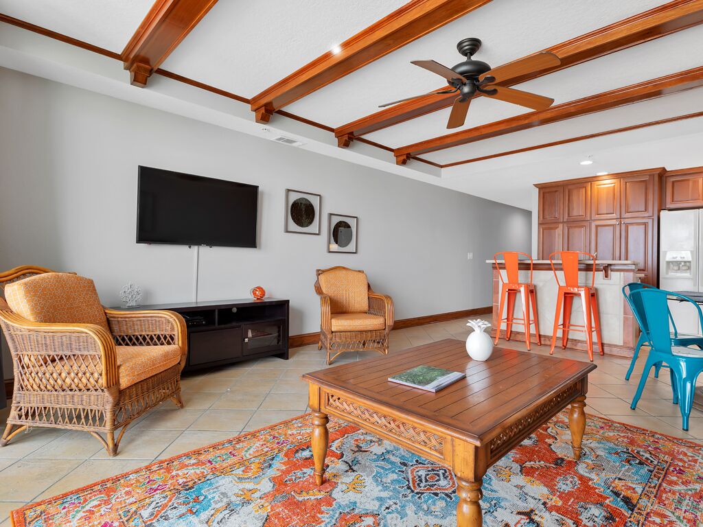 A living room with a wooden ceiling, a flat-screen TV, wicker chairs, a coffee table, and a colorful rug.