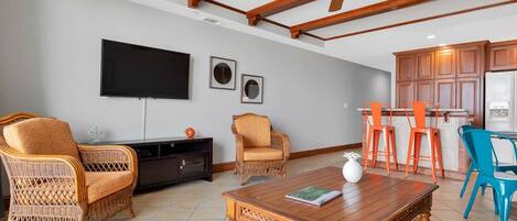 A living room with a wooden ceiling, a flat-screen TV, wicker chairs, a coffee table, and a colorful rug.