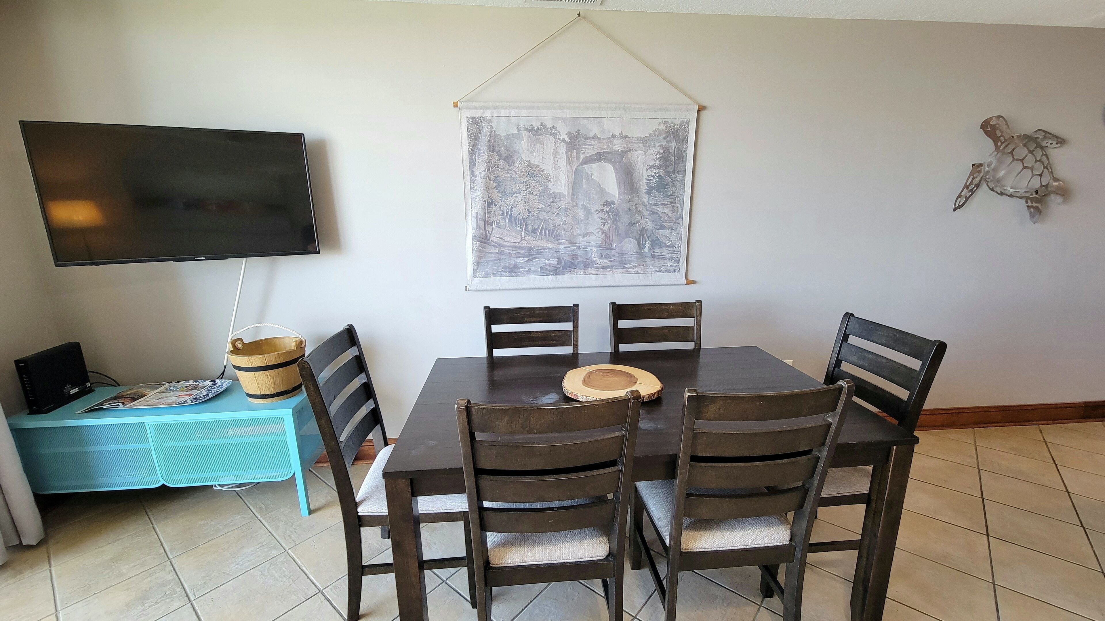 A dining area with a table, chairs, a television, and a painting on the wall.