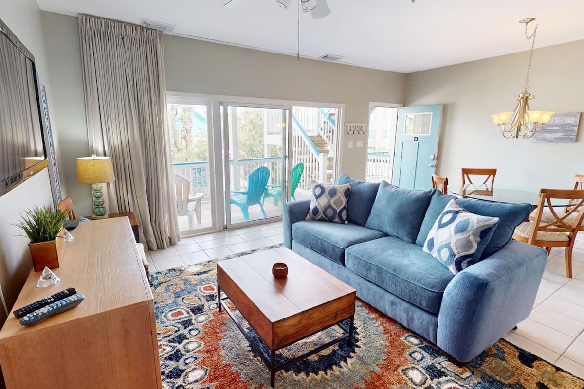 A living room with a sofa, coffee table, and a TV. There is a sliding glass door leading to a balcony with outdoor furniture.
