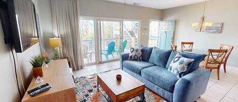 Un salon comprenant un canapé, une table basse et un téléviseur. Une porte-fenêtre coulissante donne accès à un balcon aménagé avec du mobilier d’extérieur.