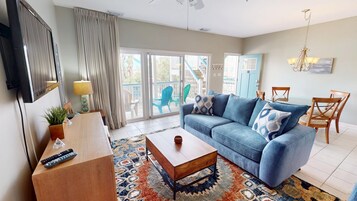 A living room with a sofa, coffee table, and a TV. There is a sliding glass door leading to a balcony with outdoor furniture.