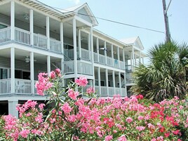 A multi-story building with balconies and a garden with pink flowers in the foreground.