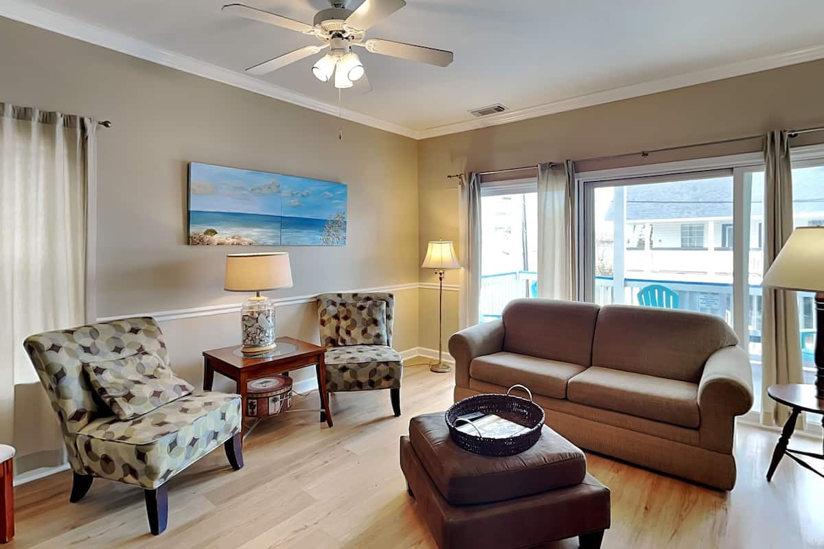 A living room with a beige sofa, patterned armchairs, a coffee table, and a ceiling fan.
