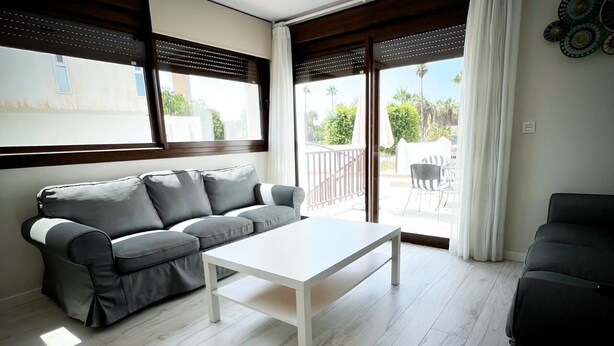 A modern living room with a grey sofa, a white coffee table, and a balcony with a view of greenery. - PUSHE Village Playa Granada Beach & Golf 3 (Motril)
