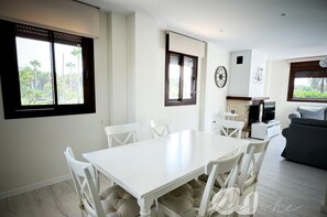 Een moderne woonkamer met een witte eettafel en stoelen, een grijze bank en een televisie.