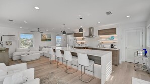 A modern kitchen with a central island, bar stools, and a dining area with a white sofa. - Gulf Front With Private Pool- Emerald View on 30A by Royal Destinations (Santa Rosa Beach)