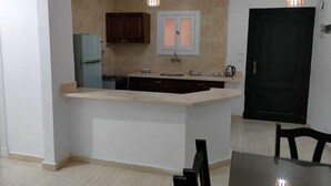 A compact kitchen with a central island, two pendant lights, a refrigerator, and a door. - Marseilia Land (El Alamein)
