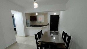 A dining area with a long table and chairs, a kitchen in the background, and a hanging light fixture. - Marseilia Land (El Alamein)