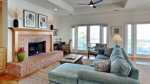 A spacious living room with a brick fireplace, a large grey sofa, and a ceiling fan. - 5 Oclock Somewhere by Tybee Vacation Rentals (Tybee Island)