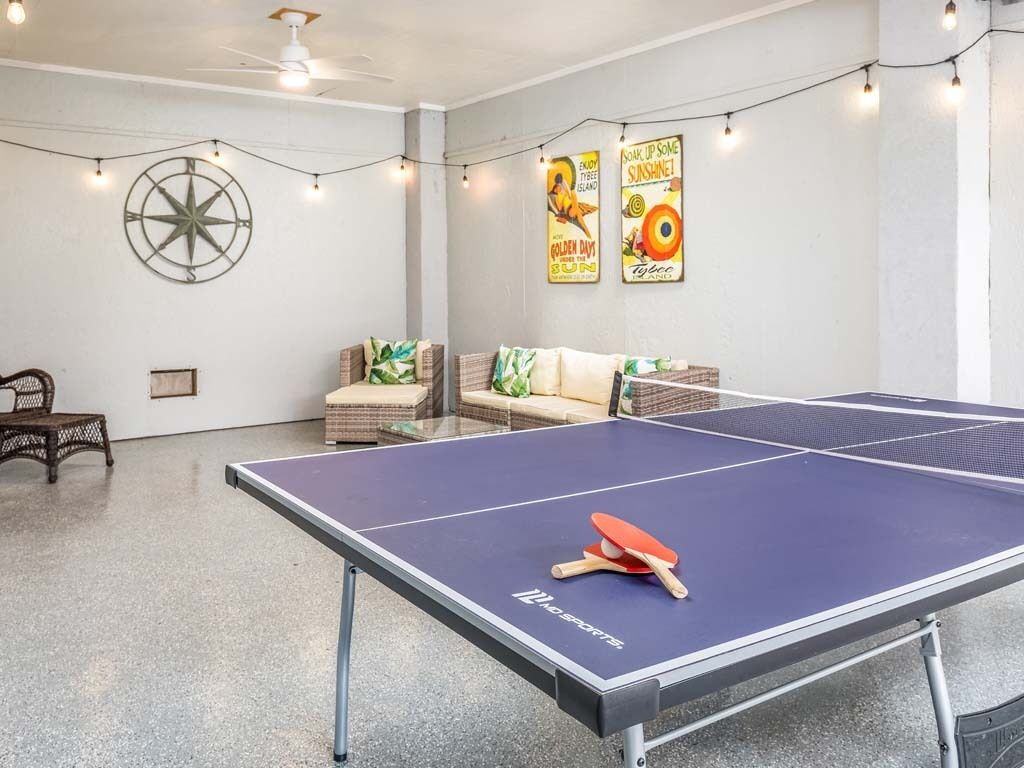 Une table de ping-pong avec une raquette et une balle, une chaise en osier, un canapé et des guirlandes lumineuses dans une pièce décorée d’une boussole au mur et de quelques affiches.