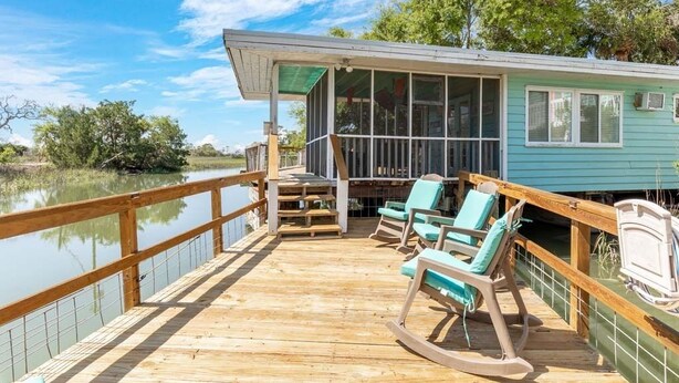 A wooden deck with rocking chairs and a covered seating area, overlooking a body of water and a building with large windows. - All Decked Out by Tybee Vacation Rentals (Tybee Island)