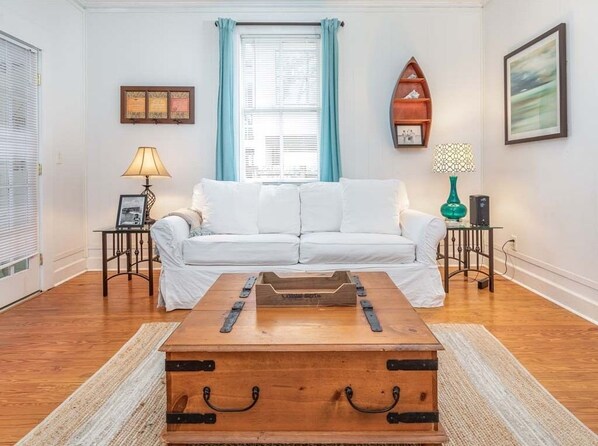 A living room with a white sofa, a wooden coffee table, a small side table with a lamp, a framed certificate on the wall, and a picture frame.