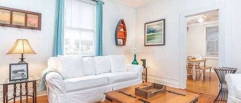 Una sala de estar con un sofá blanco, una mesa de centro de madera, una mesita auxiliar y un certificado enmarcado en la pared.