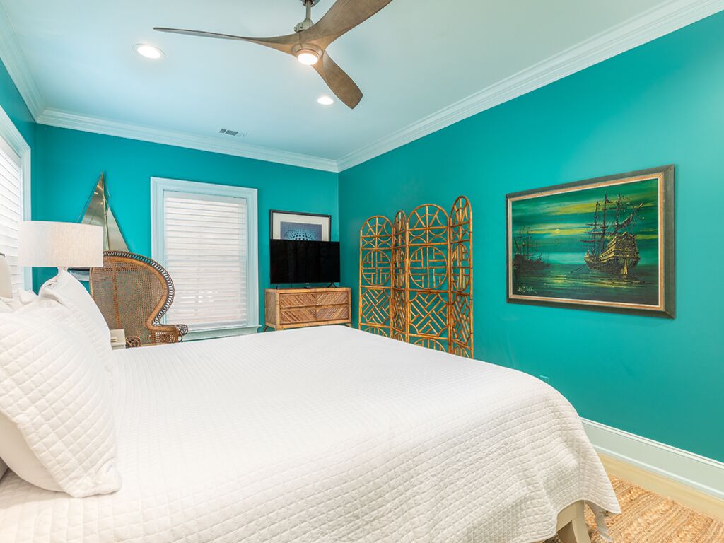 Une chambre avec des murs de couleur turquoise, un lit avec une literie blanche, un ventilateur de plafond, une télévision, un paravent décoratif et un tableau encadré.