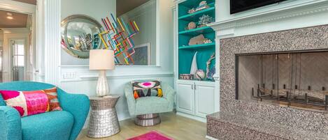 A living room with teal armchairs, a fireplace, a television, and decorative items on shelves.