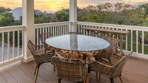 A covered outdoor patio with a round glass table and wicker chairs, surrounded by a white railing, overlooking a landscape with trees and a sunset. - Aqua Vitae by Tybee Vacation Rentals (Tybee Island)