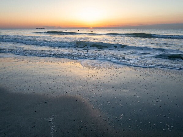 Una playa al atardecer con olas, un barco y una persona practicando surf a lo lejos.
