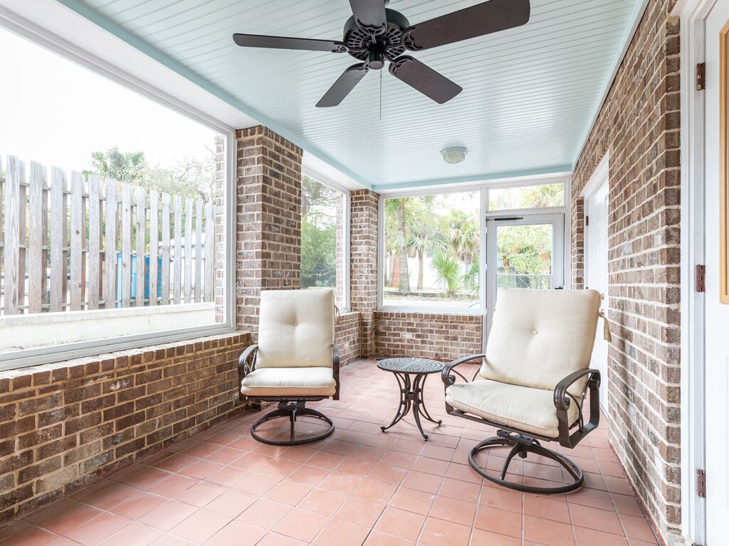 A covered patio with two cushioned chairs and a small table, brick walls, and a ceiling fan.