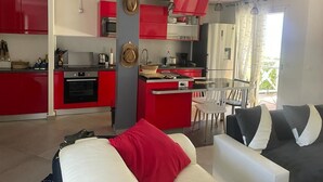 Una cuina moderna amb armaris vermells, una zona de menjador amb taula i cadires blanques, i un saló amb sofà i coixins.