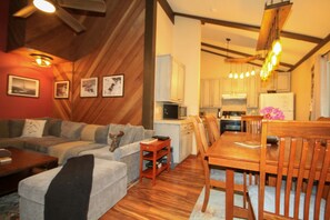 A cozy living room with a sofa, coffee table, and a dining area with a wooden table and chairs. A kitchen with cabinets and a microwave is visible in the background.