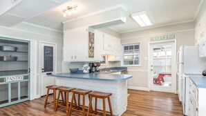 A kitchen with a central island, wooden stools, white cabinets, and a view of the outside. - Beach Bum Inn by Tybee Vacation Rentals (Tybee Island)