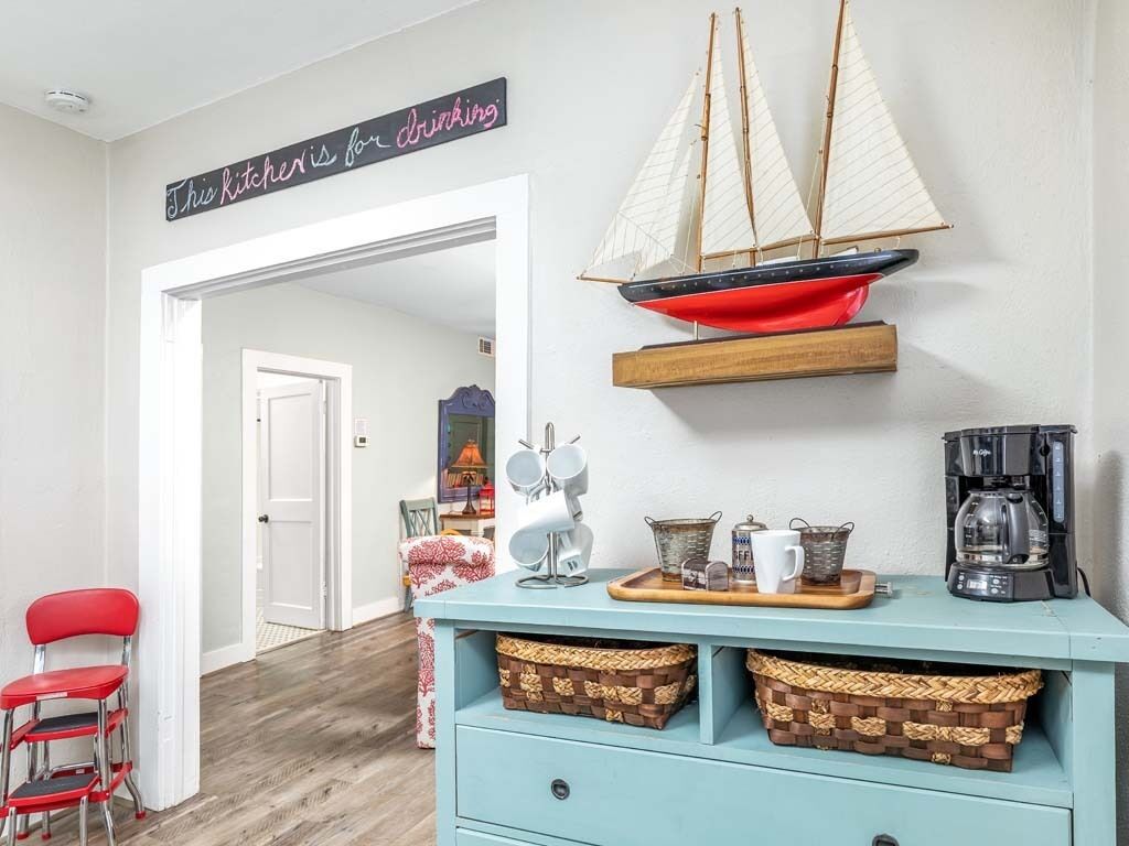 A kitchen with a sign reading "This kitchen is for drinking," a sailboat decoration, a coffee maker, and wicker baskets on a light blue cabinet.