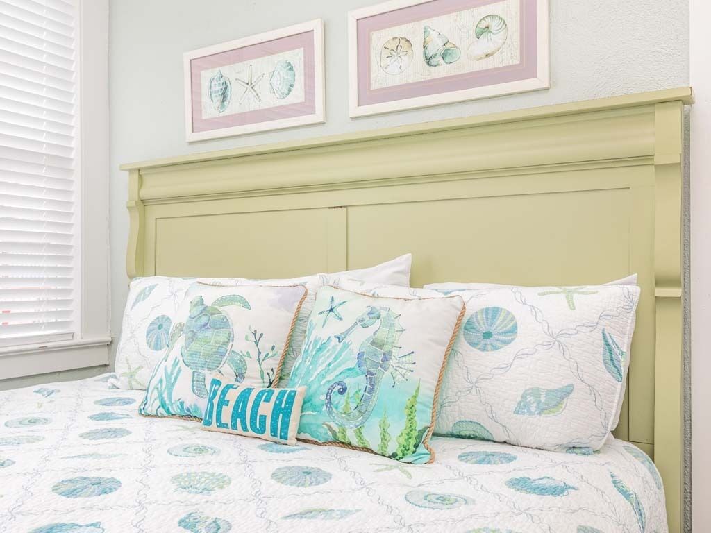 Une chambre à coucher avec un lit agrémenté d’une couvre-lit et de coussins sur le thème de la plage, d’une tête de lit vert clair et d’œuvres d’art encadrées représentant des paysages marins accrochées au mur.
