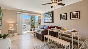 A modern living room with a sofa, coffee table, and dining table. There is a ceiling fan, a large window, and a door leading to a patio. - Spacious 3BR-  Yard - BBQ - Parking! (Costa Mesa)