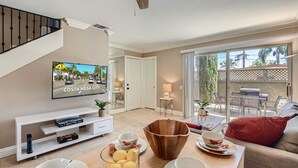 A modern living room with a flat-screen TV, a wooden coffee table, and a dining area with a red cushion. - Spacious 3BR-  Yard - BBQ - Parking! (Costa Mesa)