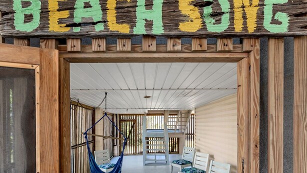 Une structure en bois sur laquelle on a peint les mots « BEACH SONG ». À l’intérieur, on trouve deux chaises blanches avec des coussins à motifs, un hamac bleu et un banc en bois.