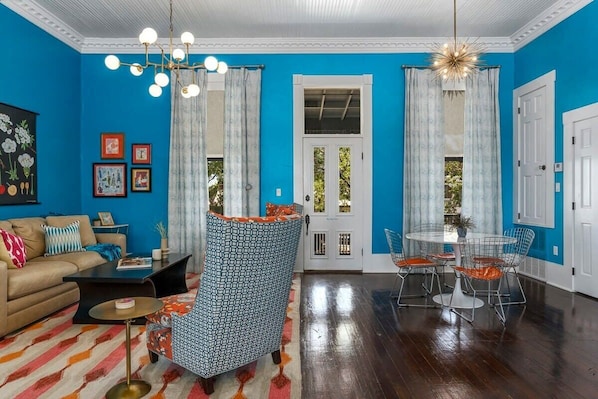 A living room with a blue wall, a sofa, a coffee table, a patterned armchair, and a dining area with a table and chairs. - The Loft on Main St, Balcony Views, Roman bathtub! (Fredericksburg)